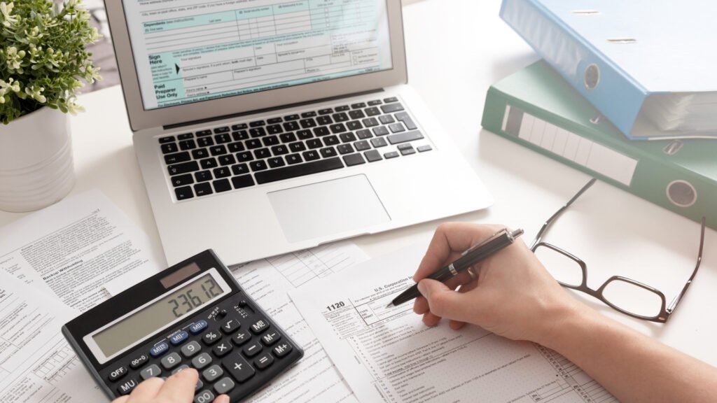 imagem de mesa com notebook, livros, calculadora e mão que escreve em papel
