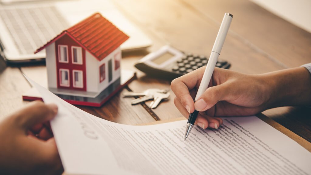 mãos assinam contrato em mesa que tem calculadora, chave e miniatura de imóvel