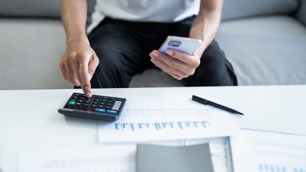 homem manuseia calculadora e papéis apoiados em uma mesa, além de segurar um celular