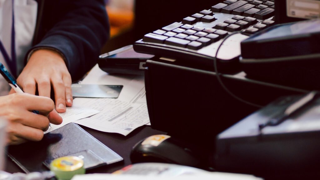 Pessoa escreve em papel numa mesa com folhas de papel, cartão de crédito e computador