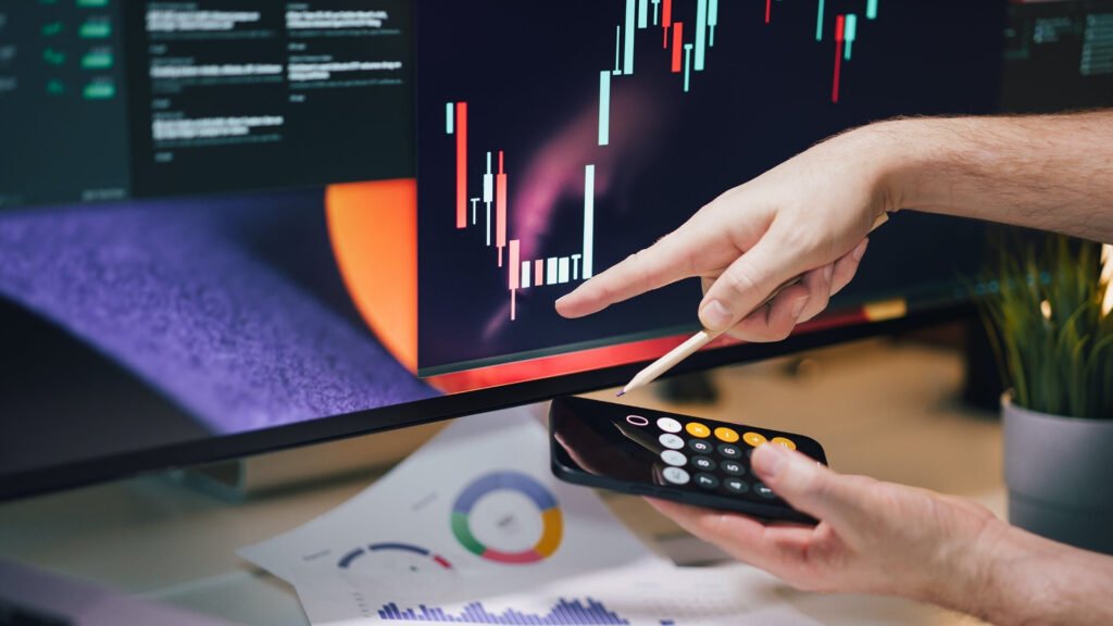 em mesa há papeis com gráficos e um computador com gráficos e uma mão aponta para tela e outra mão segura um celular com calculadora