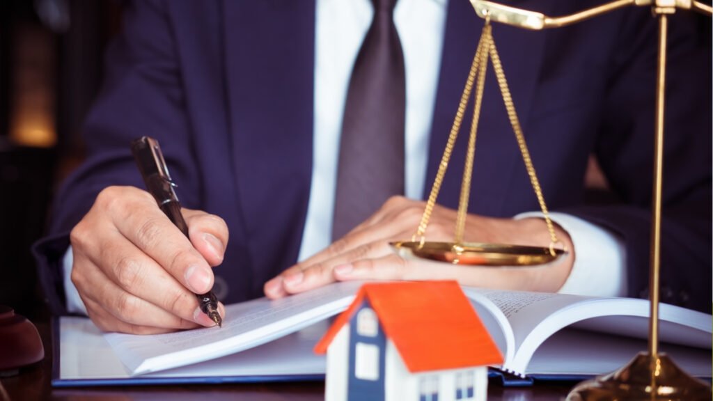 imagem contém homem de terno assinando em papel e na mesa tem balança da justiça e miniatura de casa
