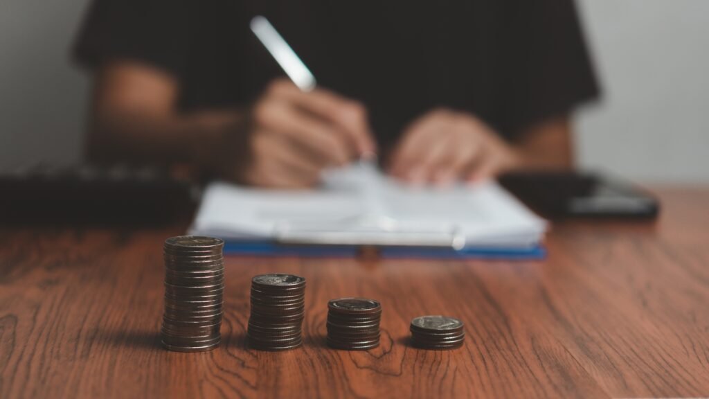 Imagem de mesa com moedas empilhadas e pessoa ao fundo escrevendo em papel